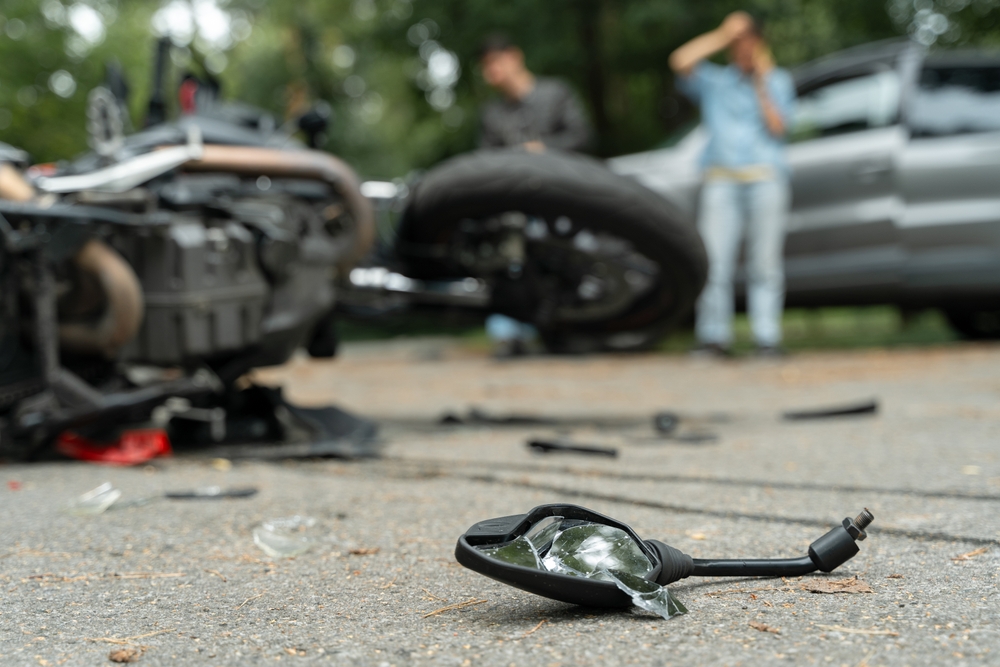 Broken motorcycle parts scattered on the road after a crash, with two people standing near the damaged vehicles